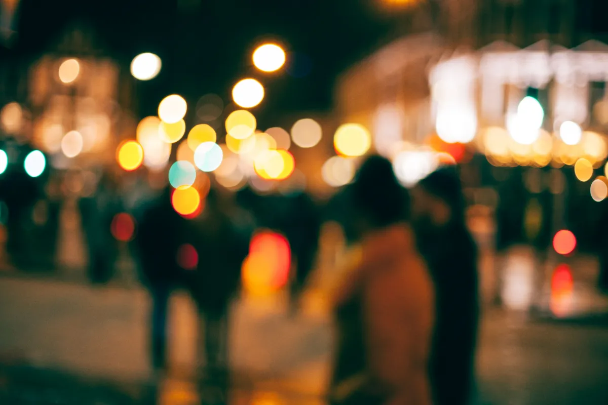 Blurred night-time city street scene with warm bokeh lights and pedestrians walking past storefronts